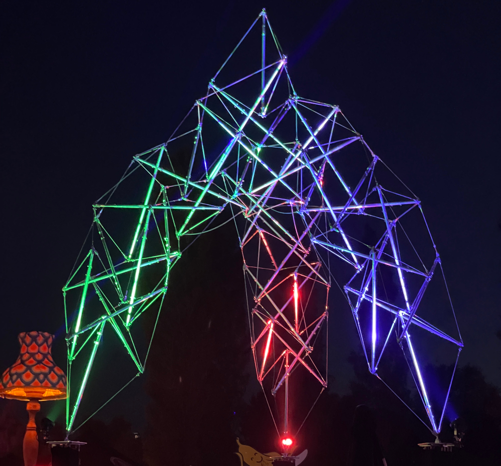 The Open Claw — a tensegrity arch of steel tubes and cable, glowing green, red and violet against the night sky.